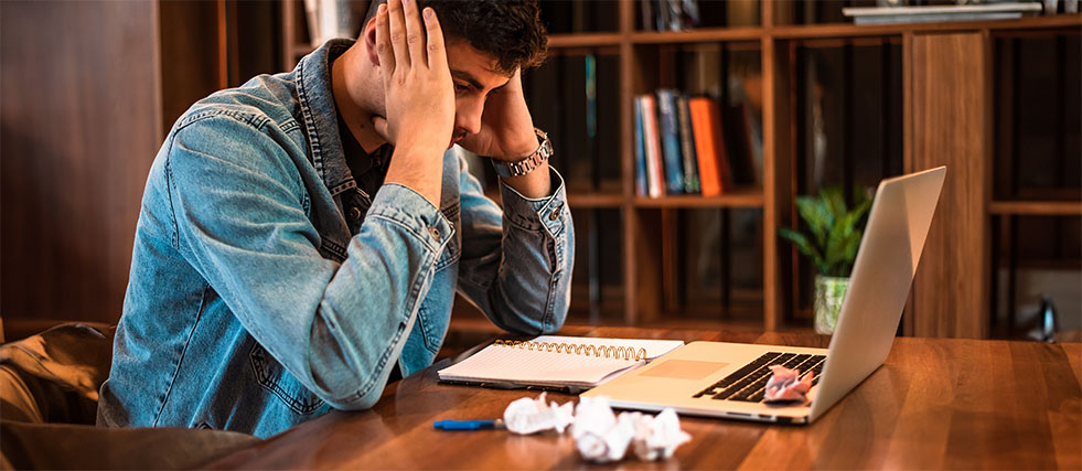 Gestreste man op werk aan bureau met laptop, notitieboek en verfrommelde papieren, opbouw naar werkgerelateerd verzuim